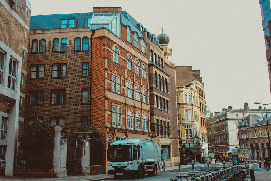 A city street scene featuring a collection of older, multi-story brick buildings with varying architectural styles, including arched and rectangular windows. A light blue refuse collection truck is pa