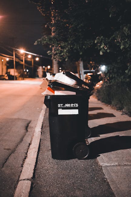 A waste collection worker, wearing a high-visibility yellow vest with reflective strips and dark clothing, is operating the controls of a large refuse collection vehicle parked on a city street during